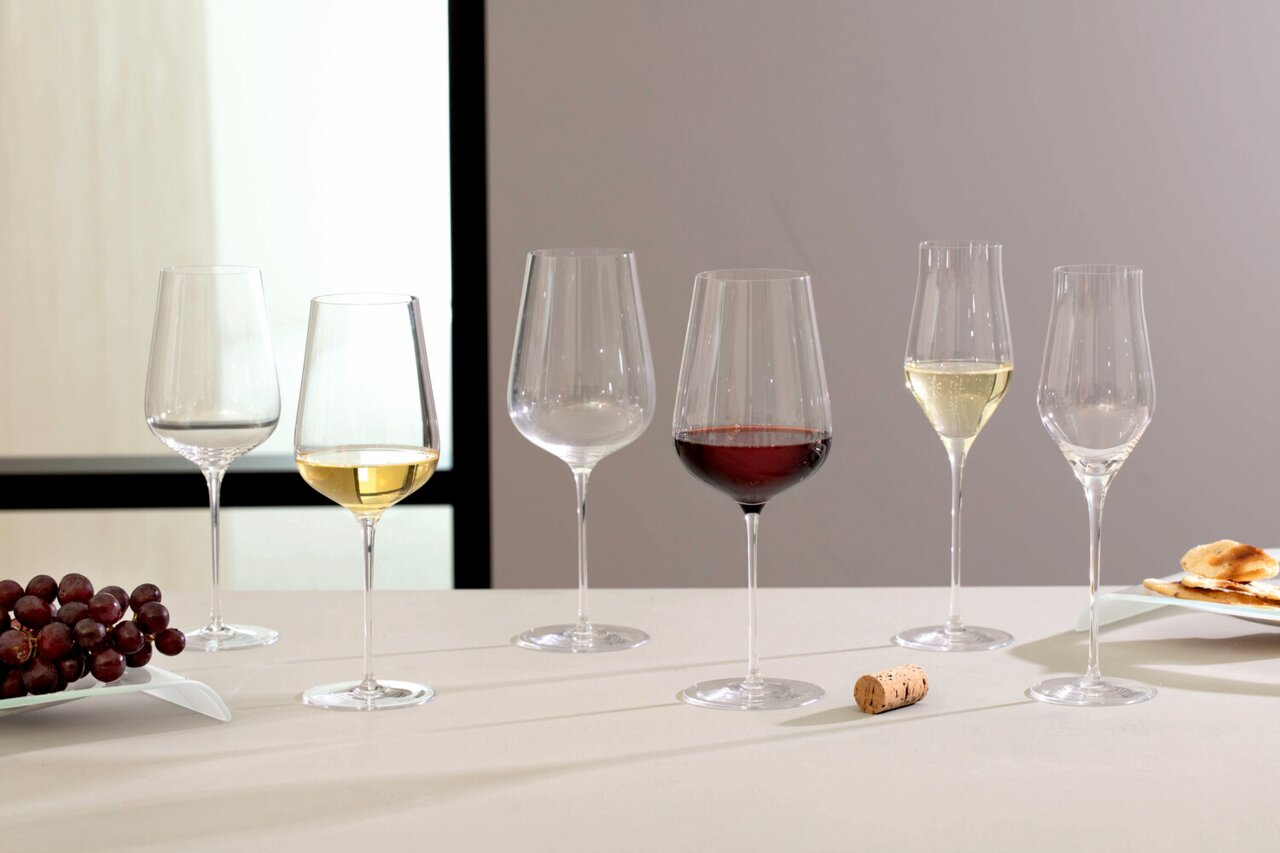 Leonardo Rodewijnglas BRUNELLI Een set van zes wijnglazen, waaronder rode- en wittewijnglazen, op een tafel met op de voorgrond een kurk en druiven. Zijaanzicht.