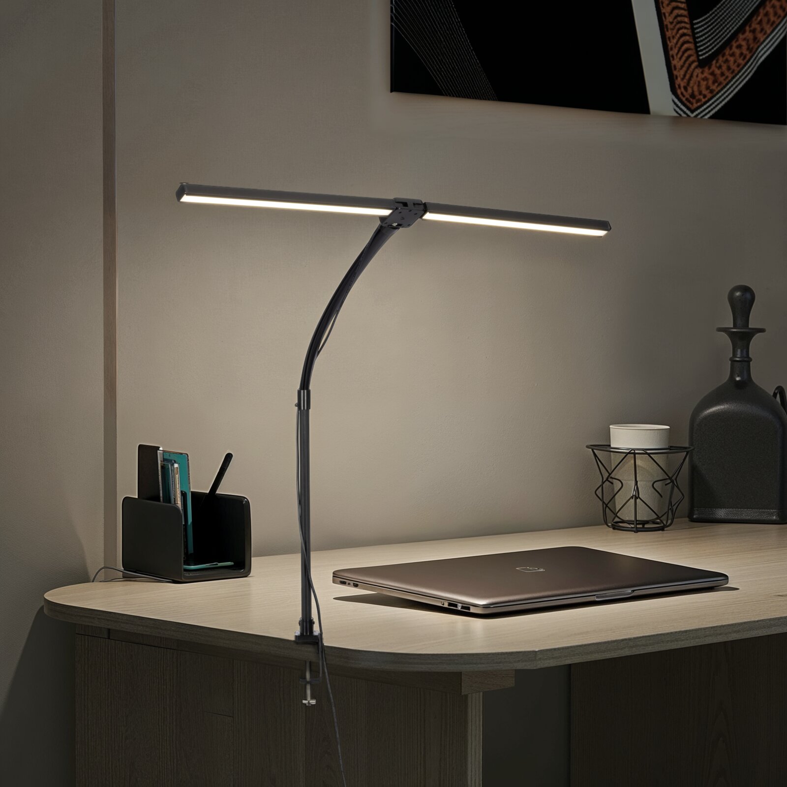 Schwarze LED-Schreibtischlampe mit Klemmbefestigung und gebogenem Arm, eingeschaltet über einem Schreibtisch mit Laptop; Perspektive: frontale leicht schräge Seitenansicht