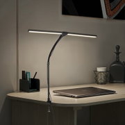 Schwarze LED-Schreibtischlampe mit Klemmbefestigung und gebogenem Arm, eingeschaltet über einem Schreibtisch mit Laptop; Perspektive: frontale leicht schräge Seitenansicht
