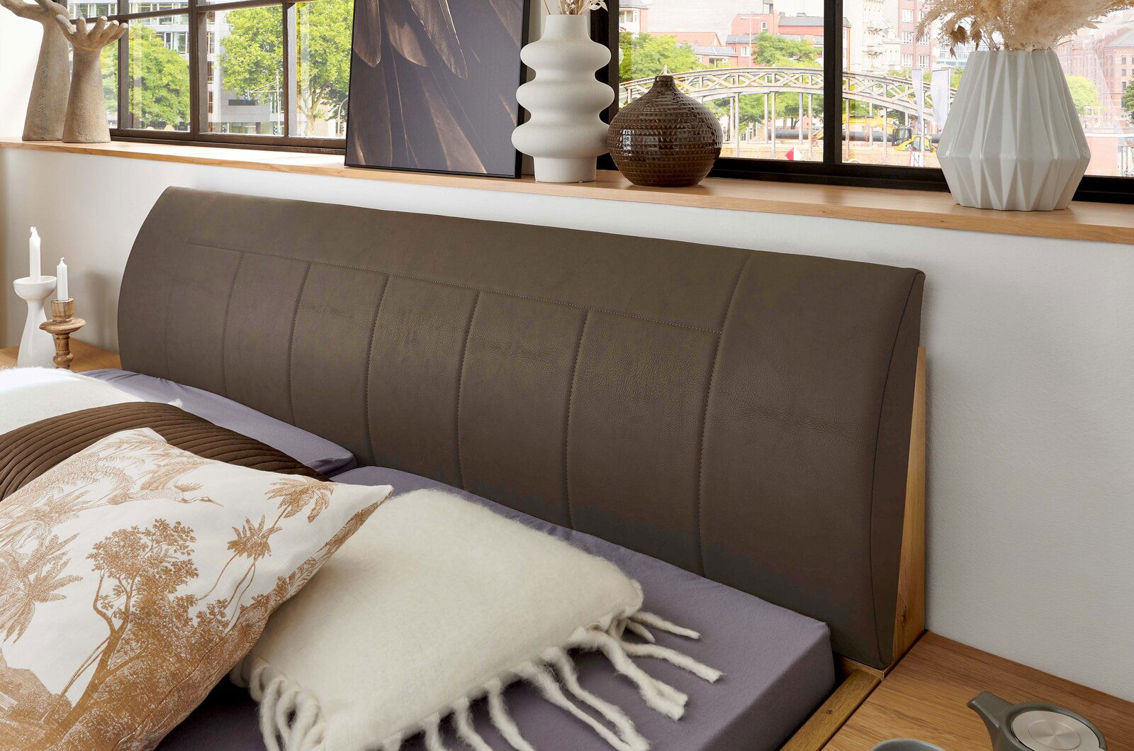 Nahaufnahme eines modernen Bettes mit gepolstertem, dunkelbraunem Kopfteil und Holzrahmen, fotografiert aus leicht schräger Frontperspektive im Schlafzimmer.