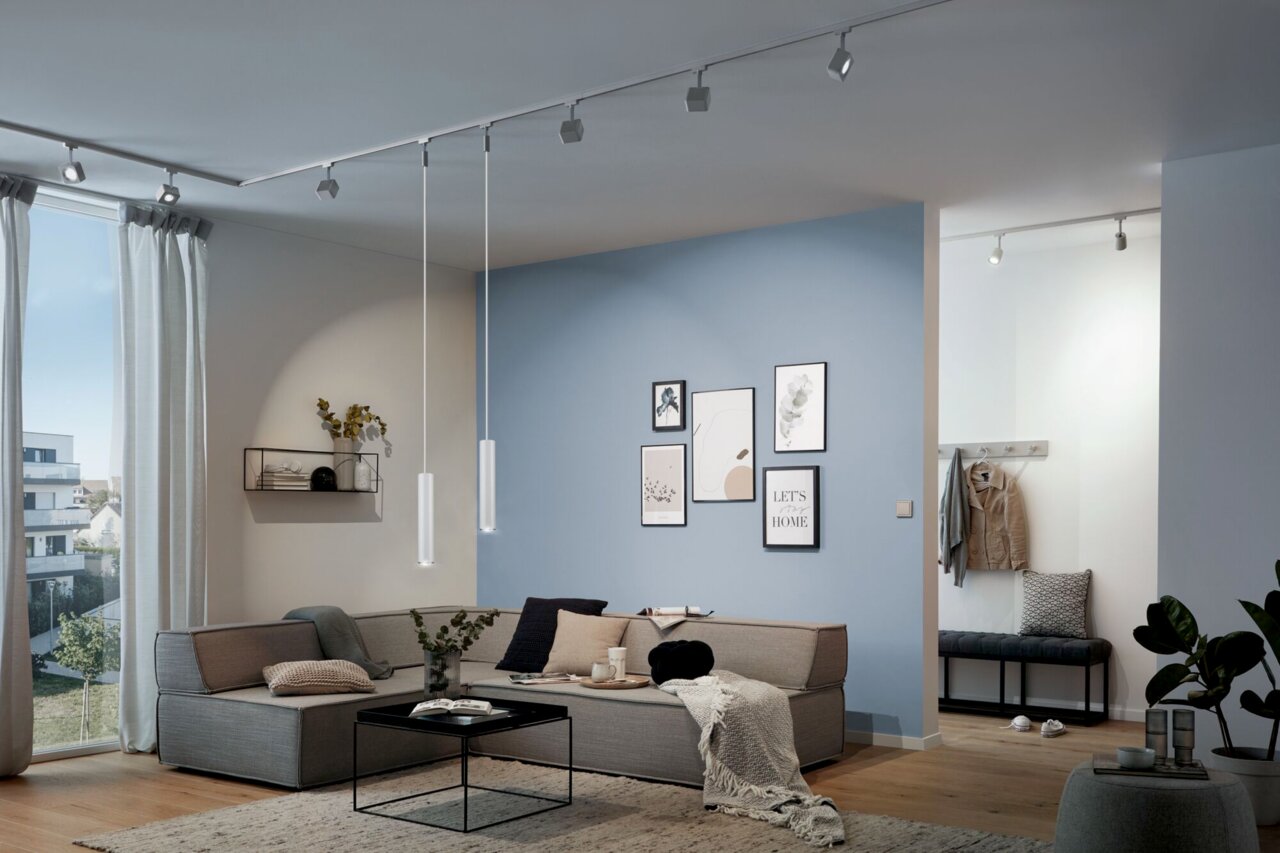 Een moderne woonkamer met de Catalejo-pendelverlichting aan een rail, die aan het plafond hangt, in een stijlvol ingerichte ruimte met een bank en wanddecoraties.