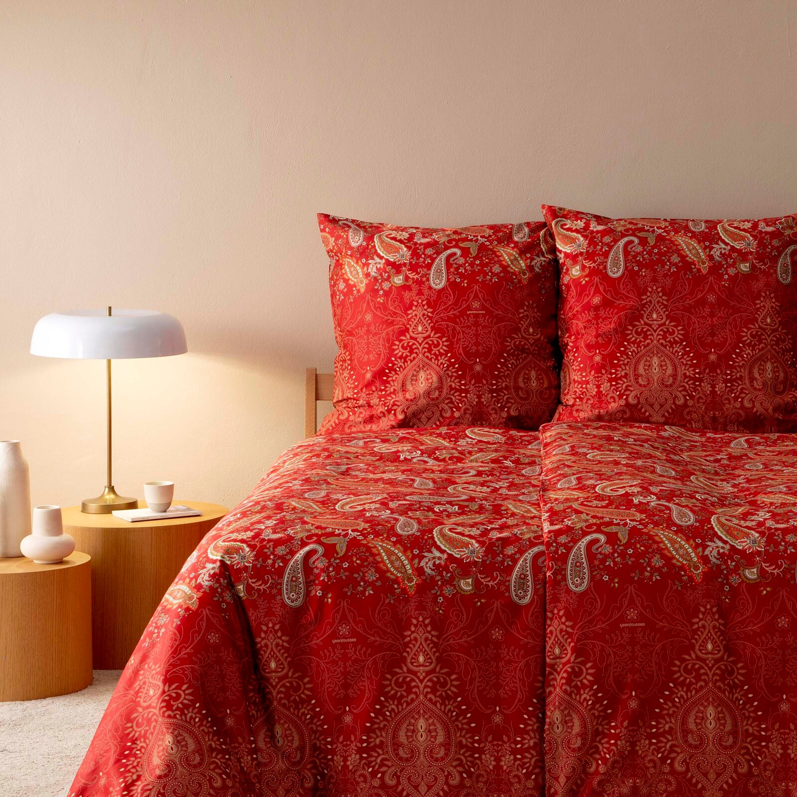 Rote Bettwäsche mit Paisley-Muster, frontal fotografiert, neben einem Nachttisch mit Lampe.