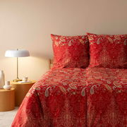 Rote Bettwäsche mit Paisley-Muster, frontal fotografiert, neben einem Nachttisch mit Lampe.