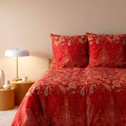 Rote Bettwäsche mit Paisley-Muster, frontal fotografiert, neben einem Nachttisch mit Lampe.