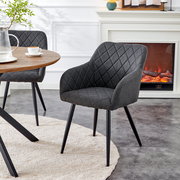 Gepolsterter Esszimmerstuhl in Dunkelgrau mit rautenförmiger Steppung und schwarzen Metallbeinen, im Wohnambiente neben Holztisch und Kamin, schräg von vorne aus leicht erhöhter Perspektive fotografiert.