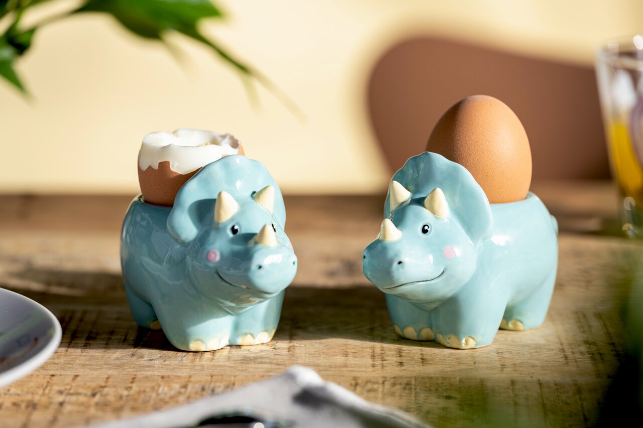 Leonardo Eierbecher Bambini Dinosaurier BAMBINI AVVENTURA Zwei Eierbecher in Form von blauen Dinosauriern auf einem Holztisch, einer mit einem gekochten Ei und der andere mit einem ganzen Ei, aus seitlicher Perspektive fotografiert.