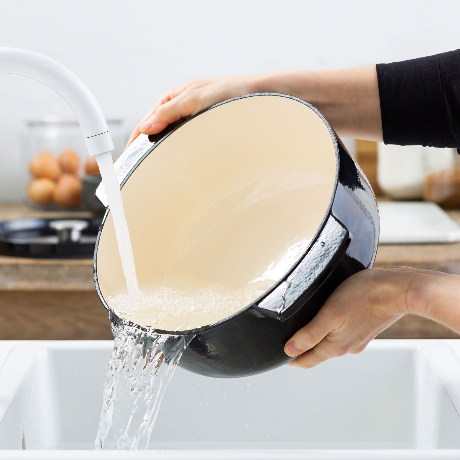 Gusseiserner Bräter rund 26cm in Schwarz, wird in der Küche unter fließendem Wasser gereinigt, seitliche Perspektive.