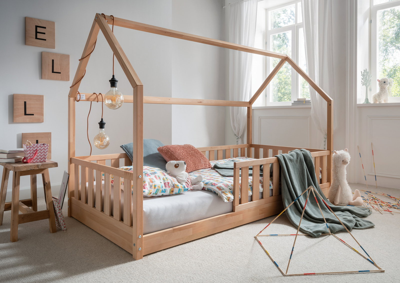 Ben 'N' Jen Hausaufsatz (für Bodenbett) INFANSKIDS Holz-Hausaufsatz für Bodenbett in einem hellen Kinderzimmer mit großen Fenstern, dekoriert mit bunten Kissen und Kuscheltieren, seitliche Perspektive.