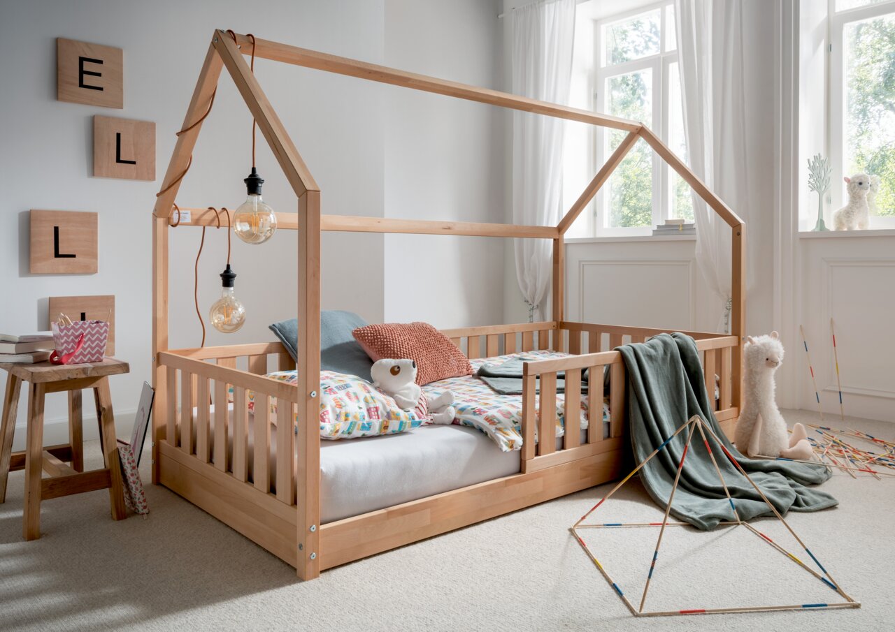 Holz-Hausaufsatz für Bodenbett in einem hellen Kinderzimmer mit großen Fenstern, dekoriert mit bunten Kissen und Kuscheltieren, seitliche Perspektive.