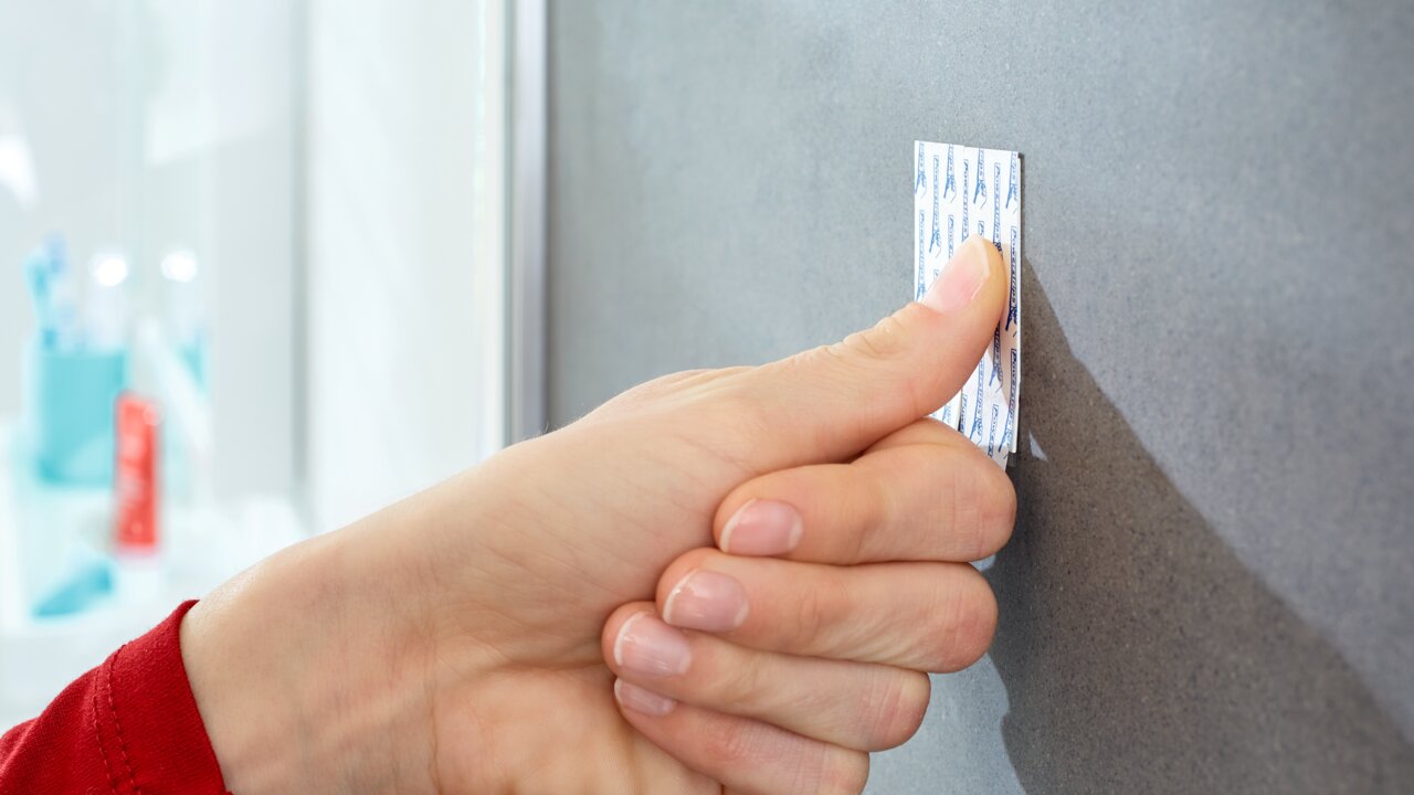 Nahaufnahme einer Hand, die einen tesa Powerstrip Waterproof an einer grauen Wand befestigt. Die Perspektive zeigt die Hand von der Seite.