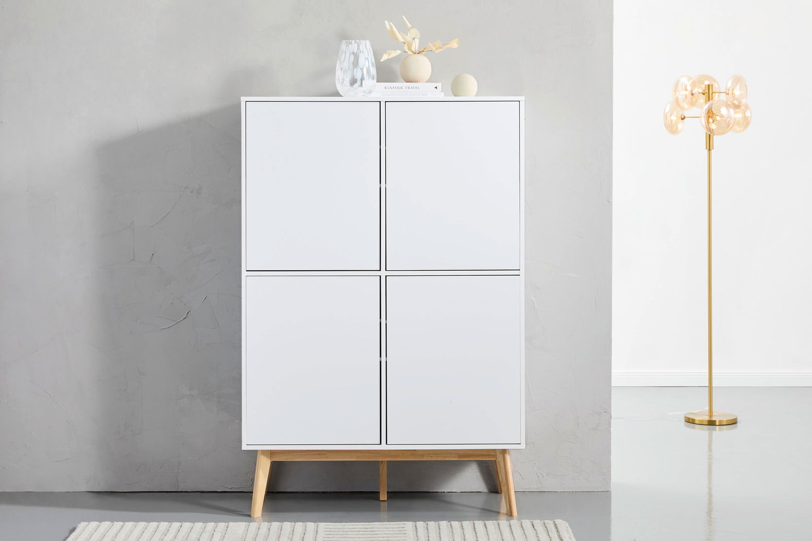 Frontale Ansicht eines modernen weißen Highboards mit vier Türen und Holzbeinen, dekoriert mit Vasen und Büchern auf der Oberseite, neben einer goldenen Stehlampe.