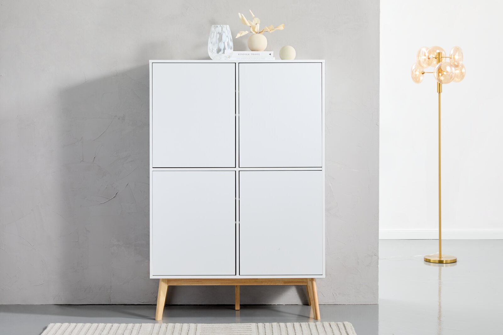 Frontale Ansicht eines modernen weißen Highboards mit vier Türen und Holzbeinen, dekoriert mit Vasen und Büchern auf der Oberseite, neben einer goldenen Stehlampe.