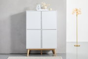 Frontale Ansicht eines modernen weißen Highboards mit vier Türen und Holzbeinen, dekoriert mit Vasen und Büchern auf der Oberseite, neben einer goldenen Stehlampe.