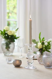 Eleganter Leuchter aus klarem Glas mit brennender Kerze, umgeben von dekorativen Vasen und Teelichthaltern auf einem Tisch. Seitliche Perspektive.