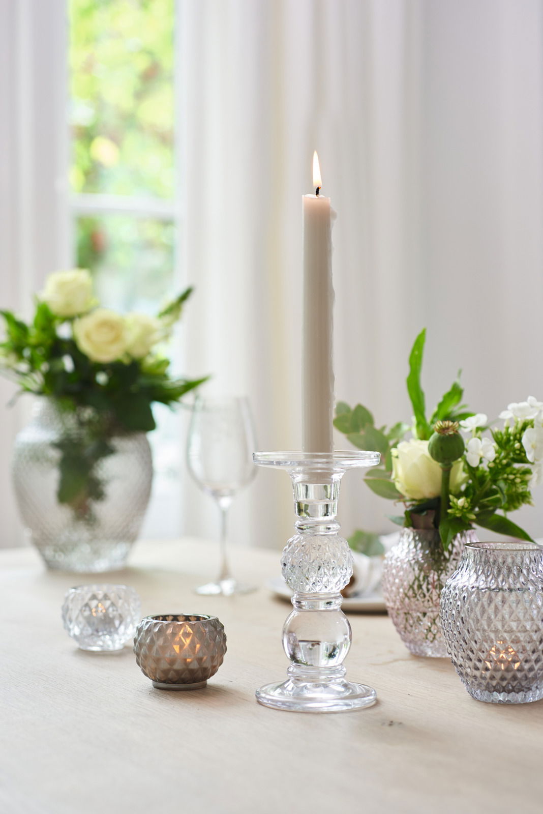Eleganter Leuchter aus klarem Glas mit brennender Kerze, umgeben von dekorativen Vasen und Teelichthaltern auf einem Tisch. Seitliche Perspektive.