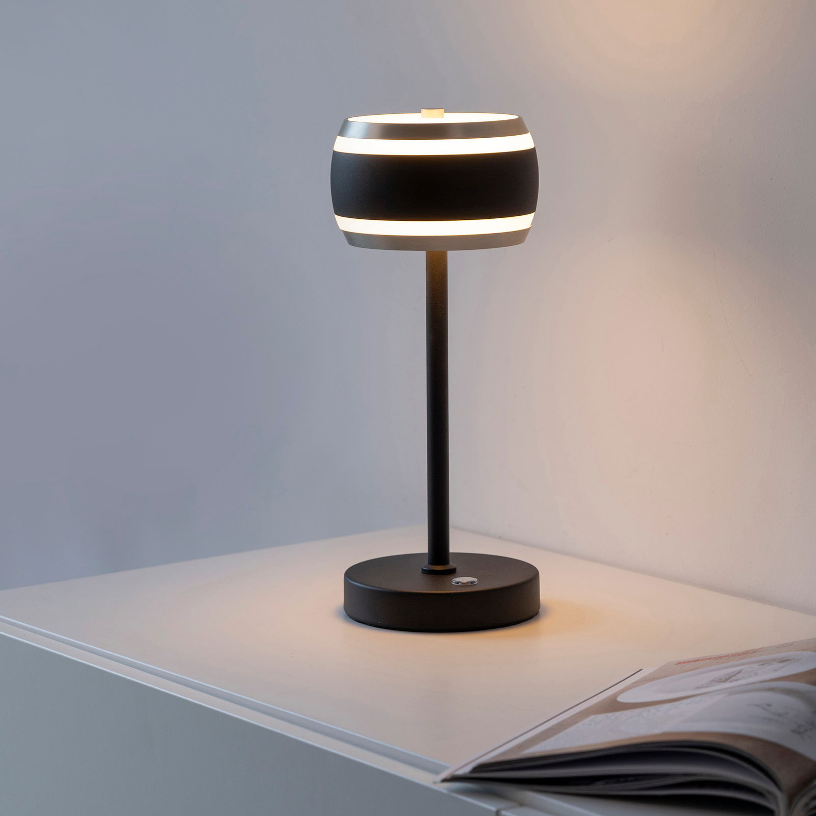 Moderne Akku-Tischleuchte in seitlicher Perspektive auf einem Tisch, mit einem runden, schwarzen Lampenschirm und warmem Licht.