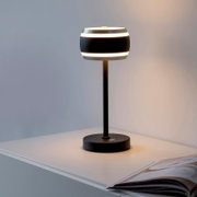 Moderne accu-tafellamp, gezien vanaf de zijkant op een tafel, met een ronde, zwarte lampenkap en warm licht.