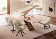 Moderner Esstisch mit Holzplatte und X-förmigem Gestell, umgeben von beigen Stühlen und einer Bank, in einem hellen Raum mit Treppe im Hintergrund, seitliche Perspektive.