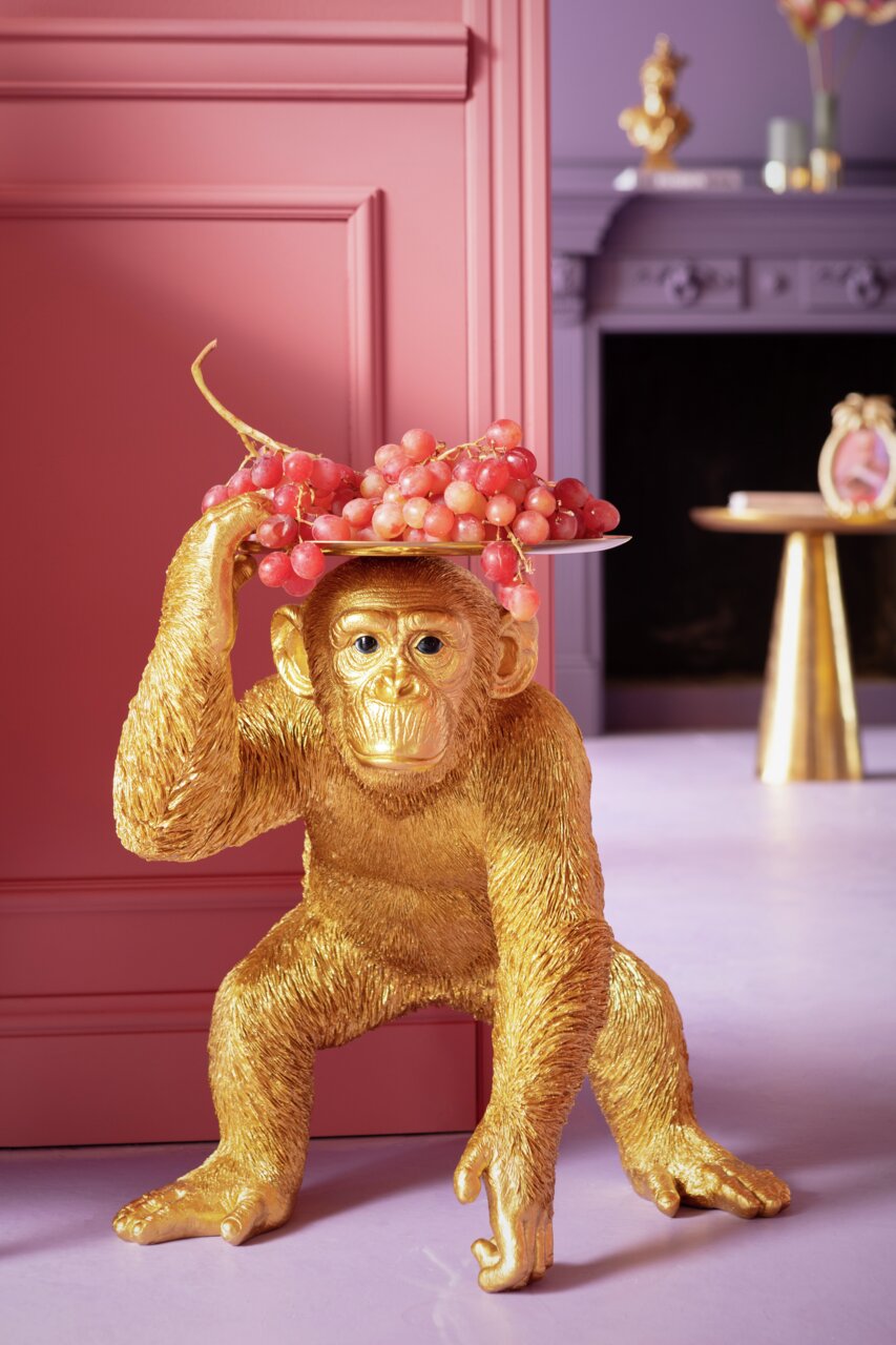 Karé Decoratiefiguur Butler Chimp Goldene Deko-Figur eines Affen in Butler-Pose, der ein Tablett mit Trauben auf dem Kopf balanciert, seitliche Perspektive.