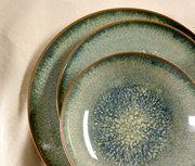 Drei übereinander gestapelte, runde Keramikteller in Blau-Grün mit gesprenkelter Glasur und braunem Rand, aus schräger Draufsicht auf hellem Stoffhintergrund.