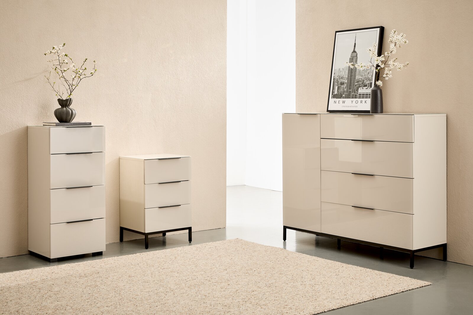 Modernes cremefarbenes Kommoden-Set mit Hochglanzfronten und schwarzen Metallgriffen sowie Füßen, bestehend aus hoher Schubladenkommode, Nachttisch und breiter Kommode, fotografiert in leicht seitlicher Frontansicht im Wohnraum-Setting