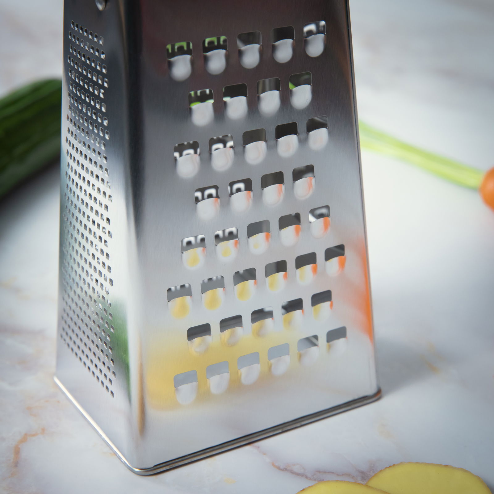 Nahaufnahme einer Vierkantreibe aus Edelstahl auf einer Küchenarbeitsplatte, mit Gemüse im Hintergrund.