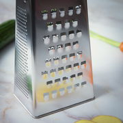 Nahaufnahme einer Vierkantreibe aus Edelstahl auf einer Küchenarbeitsplatte, mit Gemüse im Hintergrund.