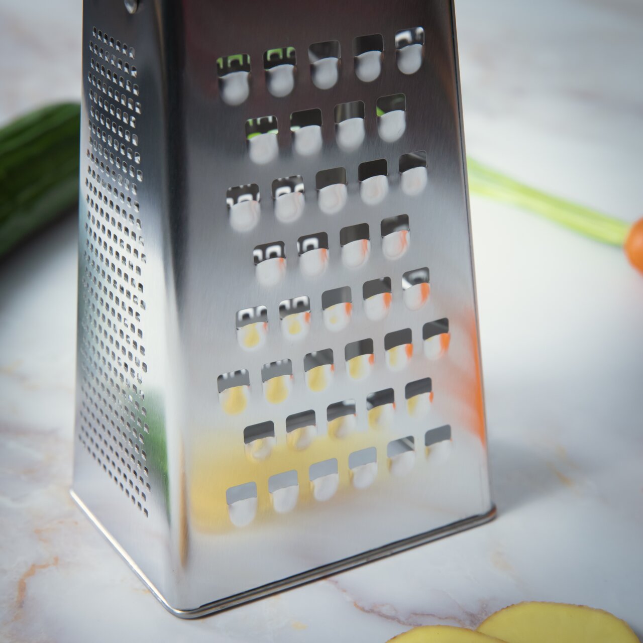 Nahaufnahme einer Vierkantreibe aus Edelstahl auf einer Küchenarbeitsplatte, mit Gemüse im Hintergrund.