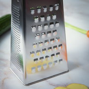 Nahaufnahme einer Vierkantreibe aus Edelstahl auf einer Küchenarbeitsplatte, mit Gemüse im Hintergrund.