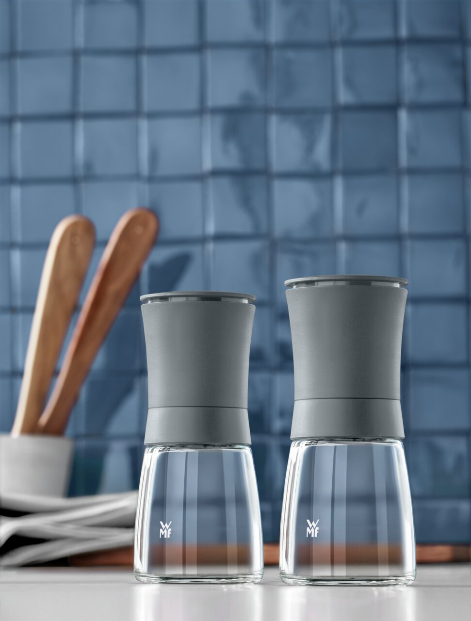 WMF Mühlenset TREND Zwei graue Mühlen aus Glas und Metall auf einer Küchenarbeitsplatte, im Hintergrund blaue Fliesen und Holzutensilien, Frontalansicht