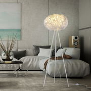 Moderne Stehleuchte LOLAsmart-NAOMI mit weißem, wolkenartigem Lampenschirm auf dreibeinigem Stativ, in einem stilvollen Wohnzimmer mit grauem Sofa und dekorativem Tisch, seitliche Perspektive.