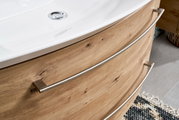 Close-up van een badkamermeubel met houtlook en gebogen metalen handgrepen, gezien vanaf de zijkant.