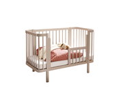 Beiges Babybett aus Holz mit abgerundeten Ecken und Gitterseiten, mit Matratze und rosa Bettdecke sowie Kuscheltier, freigestellt vor weißem Hintergrund; Perspektive: schräg von vorn (3/4-Ansicht)