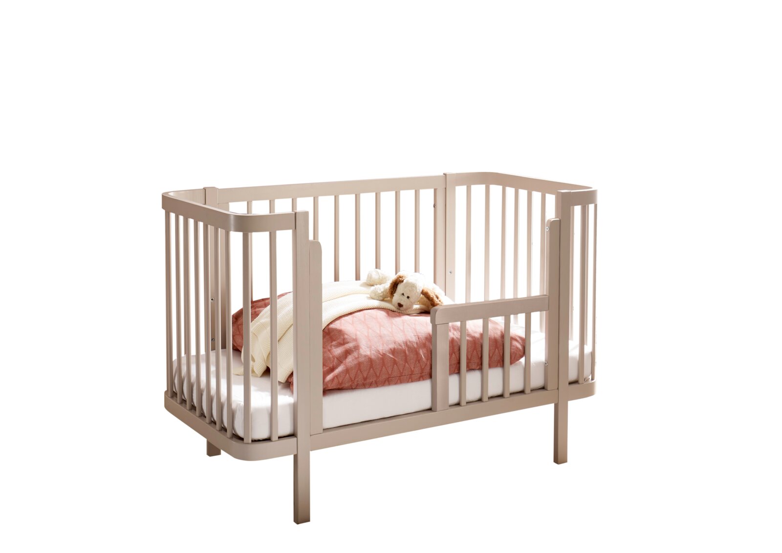 Beiges Babybett aus Holz mit abgerundeten Ecken und Gitterseiten, mit Matratze und rosa Bettdecke sowie Kuscheltier, freigestellt vor weißem Hintergrund; Perspektive: schräg von vorn (3/4-Ansicht)