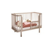 Beiges Babybett aus Holz mit abgerundeten Ecken und Gitterseiten, mit Matratze und rosa Bettdecke sowie Kuscheltier, freigestellt vor weißem Hintergrund; Perspektive: schräg von vorn (3/4-Ansicht)