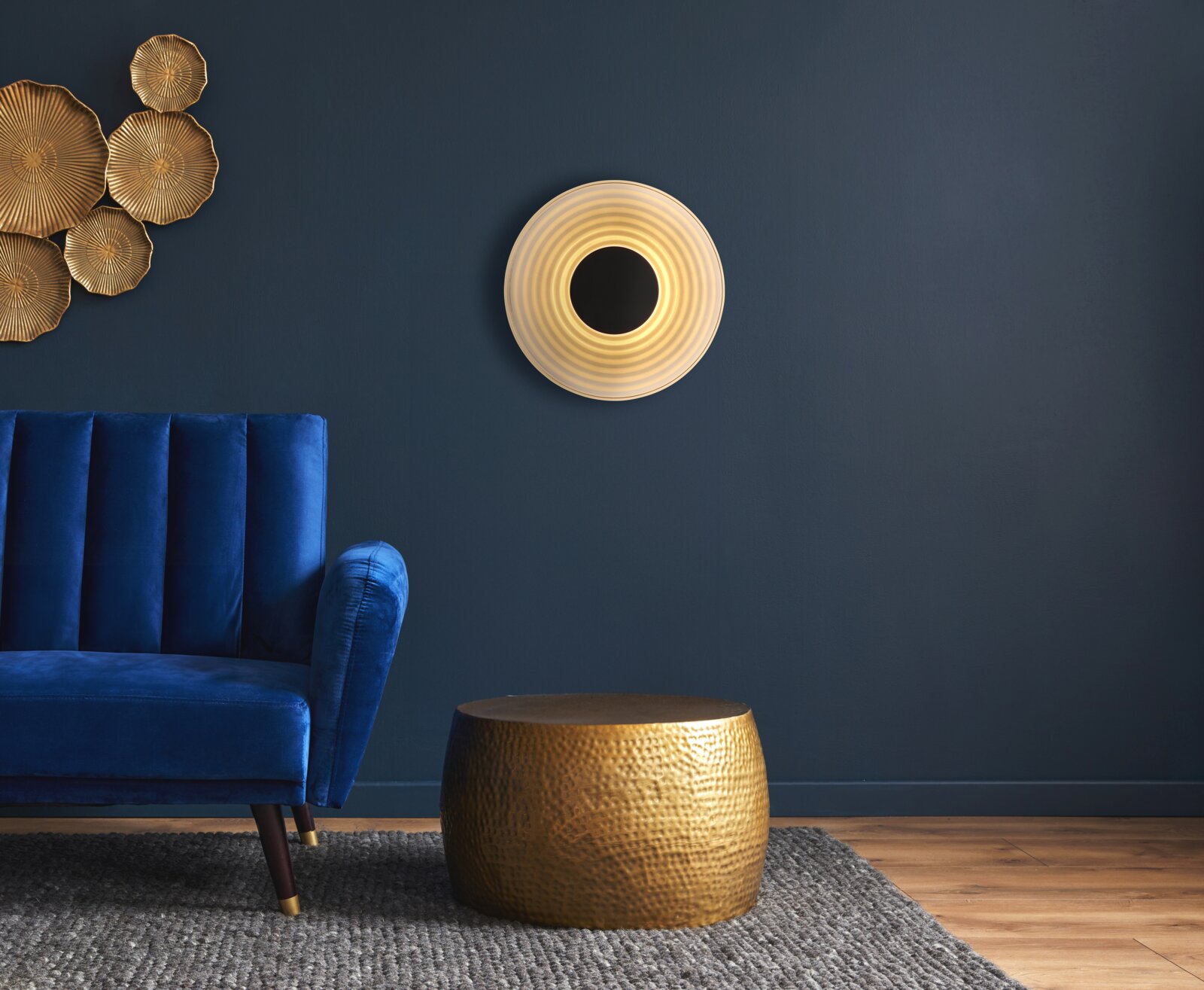 Runde, ringförmige LED-Wandleuchte mit warmweißem Licht an dunkelblauer Wand im Wohnzimmer, Frontansicht links blauer Samtsessel und goldene Wanddeko, unten goldener Beistelltisch auf grauem Teppich