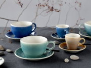 Keramik-Tassen und Espressotassen in Blau, Türkis und Beige mit passenden Untertassen auf dunkler Tischdecke, dekoriert mit Steinen und Zweigen; Perspektive: leicht von vorne in Augenhöhe mit Fokus auf der türkisfarbenen Tasse im Vordergrund