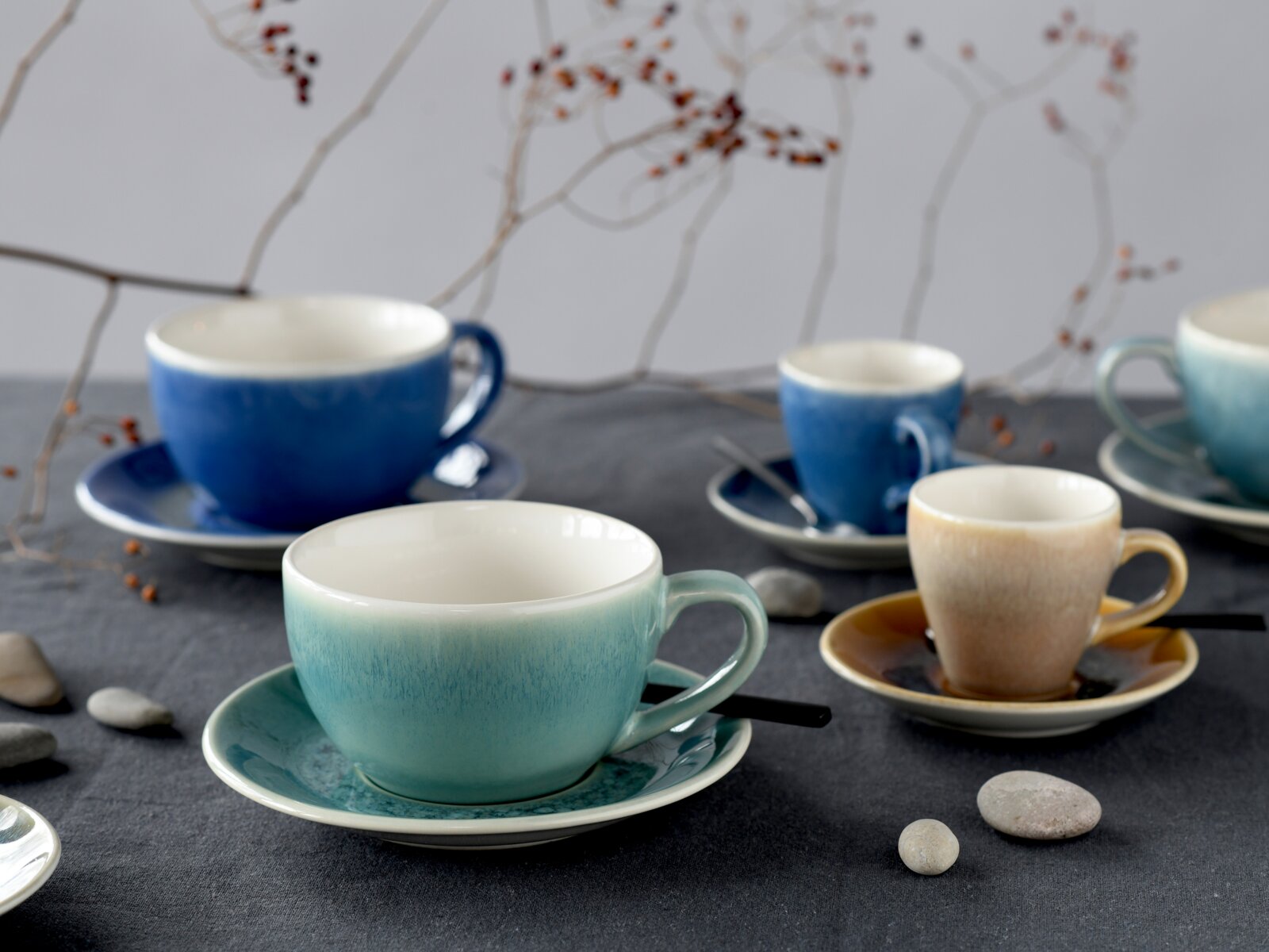 Keramik-Tassen und Espressotassen in Blau, Türkis und Beige mit passenden Untertassen auf dunkler Tischdecke, dekoriert mit Steinen und Zweigen; Perspektive: leicht von vorne in Augenhöhe mit Fokus auf der türkisfarbenen Tasse im Vordergrund