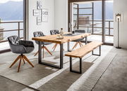 Moderner Esstisch aus Holz mit Metallbeinen, umgeben von grauen Stühlen und einer Bank, in einem hellen Raum mit Blick auf einen See. Perspektive: seitlich.