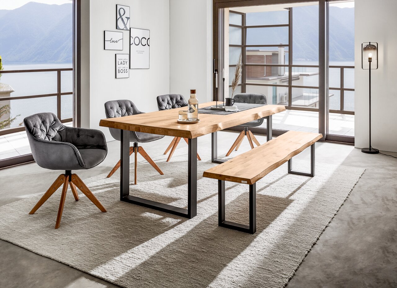 Moderner Esstisch aus Holz mit Metallbeinen, umgeben von grauen Stühlen und einer Bank, in einem hellen Raum mit Blick auf einen See. Perspektive: seitlich.
