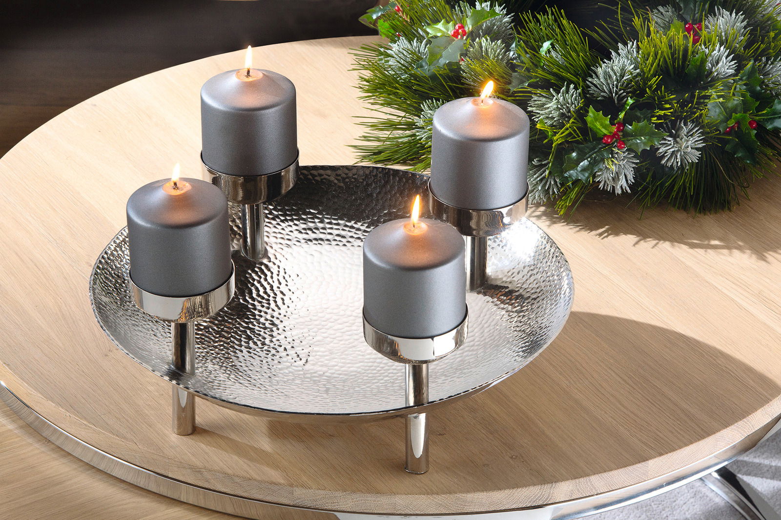 Dekorativer Kranz mit vier brennenden grauen Kerzen auf einem silbernen, gehämmerten Tablett, umgeben von grünen Tannenzweigen mit roten Beeren, aus der Vogelperspektive fotografiert.