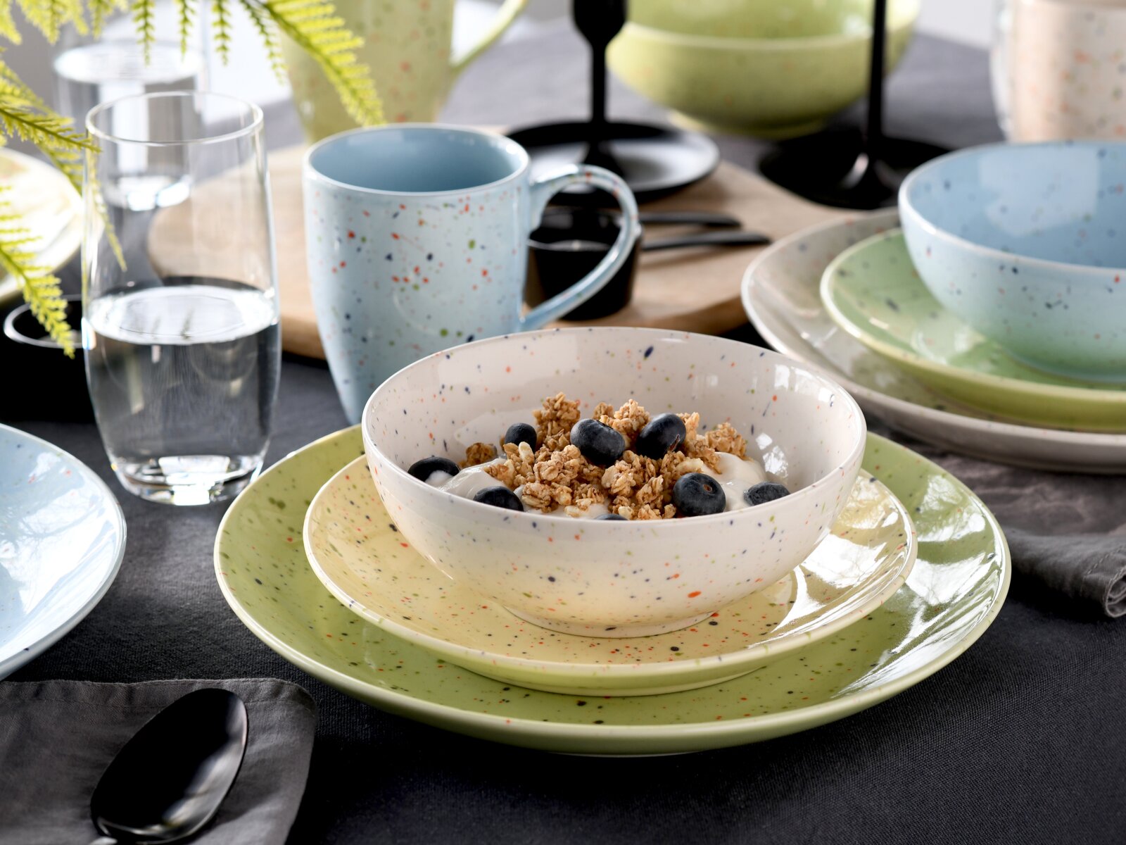 Gesprenkeltes Keramik-Geschirrset in Pastellfarben (Schale mit Müsli und Blaubeeren auf grünen Tellern, blaue Tasse und weiteres Geschirr im Hintergrund), fotografiert in leichter Schrägaufsicht von vorne.