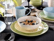 Gesprenkeltes Keramik-Geschirrset in Pastellfarben (Schale mit Müsli und Blaubeeren auf grünen Tellern, blaue Tasse und weiteres Geschirr im Hintergrund), fotografiert in leichter Schrägaufsicht von vorne.
