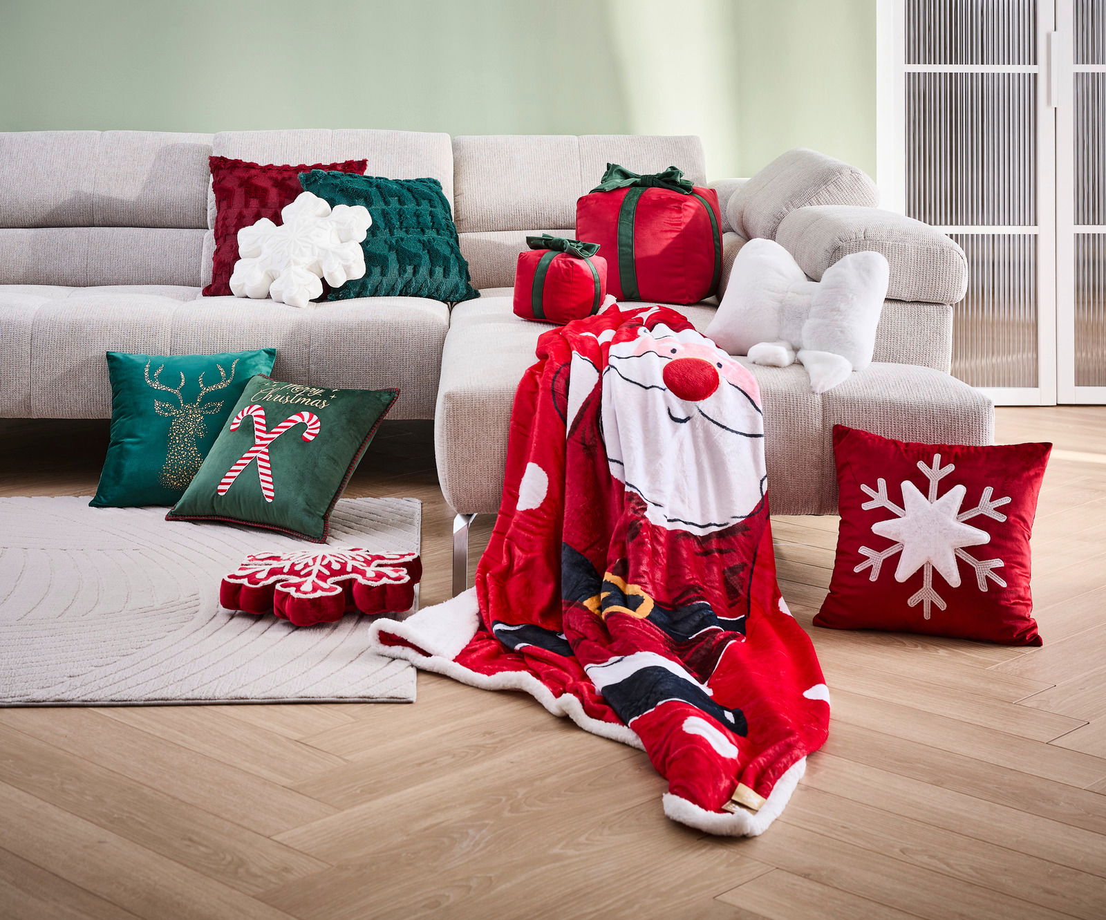 Novel Zierkissen Wohnzimmer mit mehreren dekorativen Kissen in Weihnachtsmotiven, darunter ein rotes Kissen mit Schneeflocke, ein grünes Kissen mit Rentier und Zuckerstange, sowie ein rotes Kissen in Geschenkform. Im Vordergrund liegt eine rote Decke mit Weihnachtsmannmot