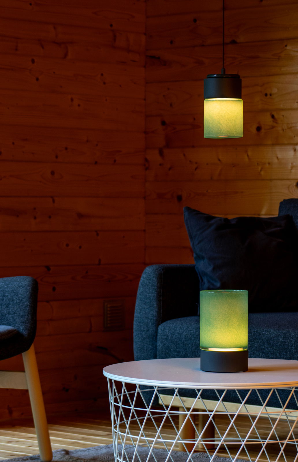 Zwei moderne, zylinderförmige Lampen mit grünlichem Licht, eine hängend und eine auf einem Tisch stehend, in einem gemütlichen Raum mit Holzwänden und einem Sofa im Hintergrund. Perspektive: seitlich.