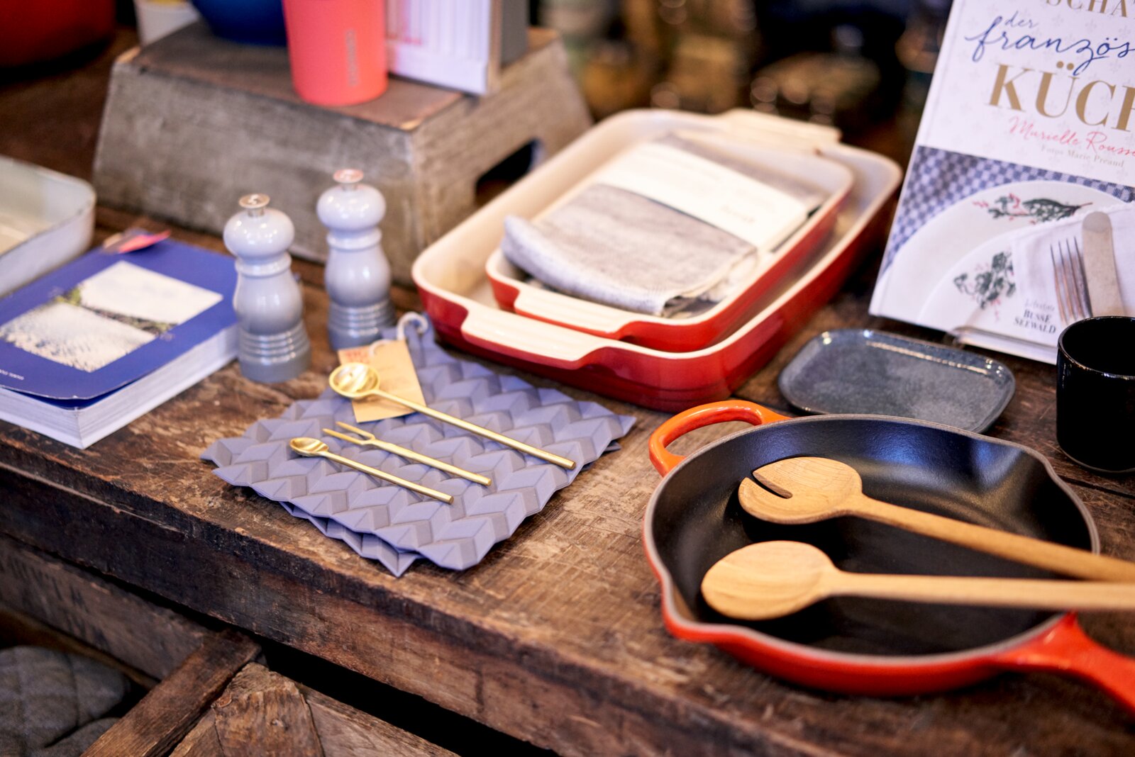 Sfeervolle collectie kook- en tafelaccessoires op een rustieke houten tafel, met rood-zwarte gietijzeren pan, houten lepels, ovenschalen, molens en goudkleurig bestek, schuin van boven gefotografeerd.