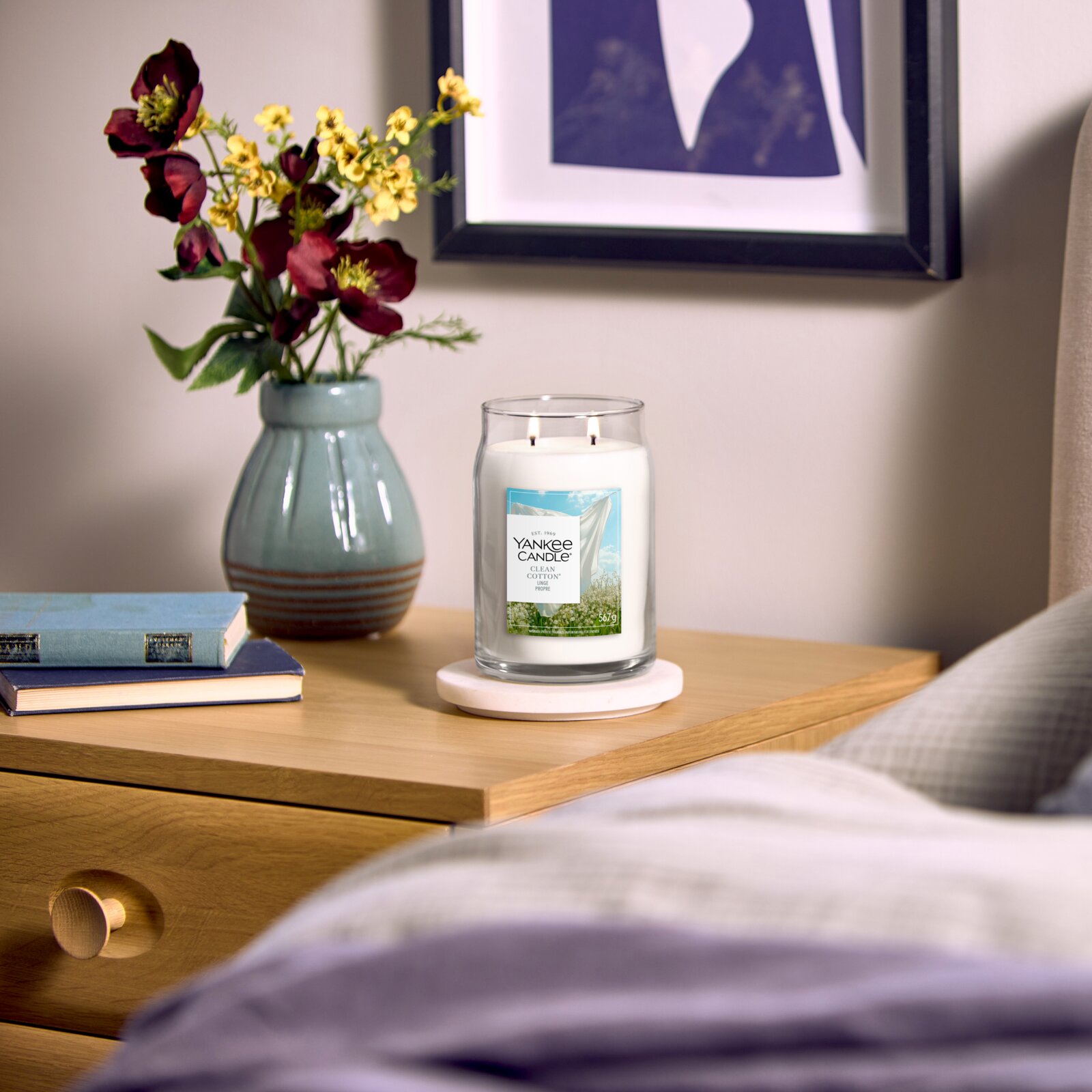 Frontalansicht einer brennenden Yankee Candle Clean Cotton Duftkerze im großen Glas mit zwei Dochten auf einem Nachttisch, stilvoll arrangiert neben Büchern und einer Blumenvase.