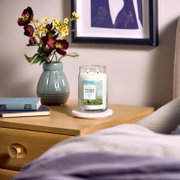 Frontalansicht einer brennenden Yankee Candle Clean Cotton Duftkerze im großen Glas mit zwei Dochten auf einem Nachttisch, stilvoll arrangiert neben Büchern und einer Blumenvase.