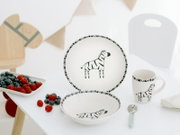 Weißes Kinder-Geschirrset mit schwarzem Zebra-Motiv, bestehend aus Teller, Schale und Henkelbecher, in leicht erhöhter Frontansicht auf einem gedeckten Tisch präsentiert