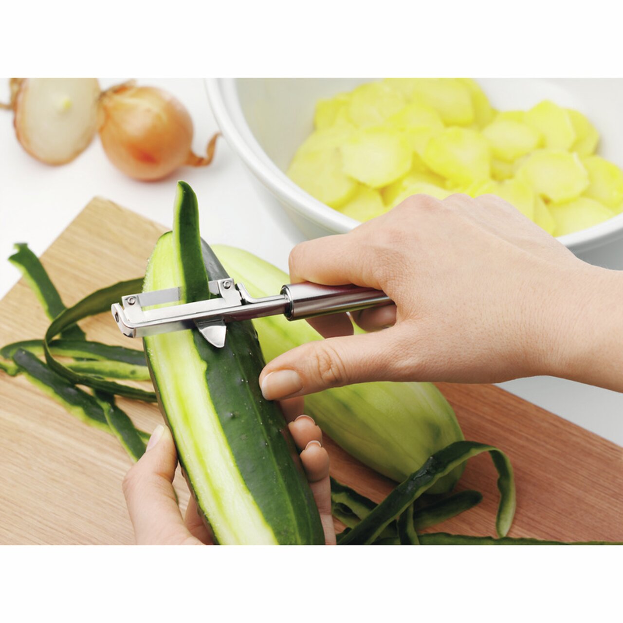 Nahaufnahme eines Pendelschälers PROFI PLUS, der eine Gurke schält. Der Schäler wird von einer Hand gehalten und zeigt die Funktionalität des Produkts.